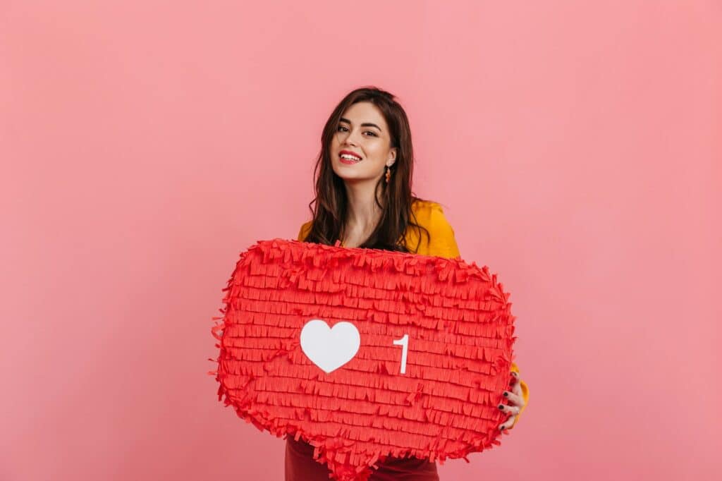 Teenager girl in bright outfit is smiling, holding red "Like" sign from Instagram on pink backgroun